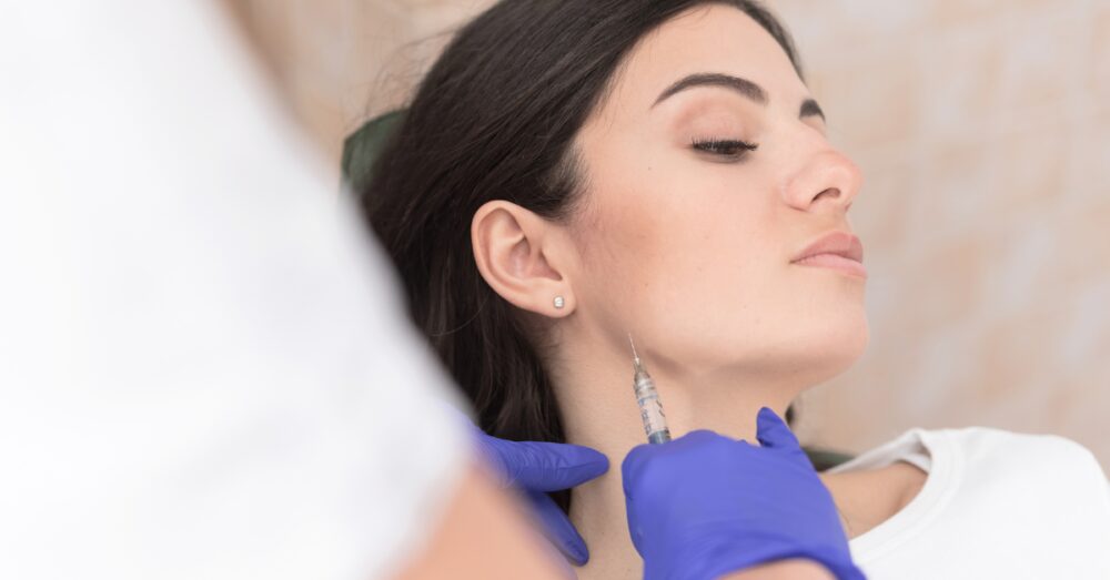 A woman with dark hair tilting her head to the side as a provider in purple gloves prepares to administer a masseter Botox injection along her jawline