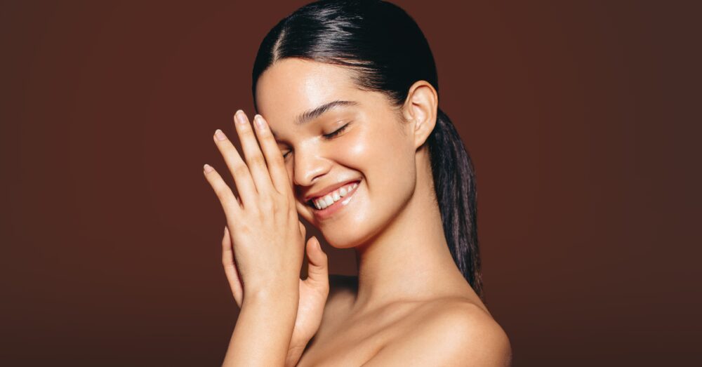 A woman with her eyes closed and hand resting near her cheek, smiling softly against a warm brown background
