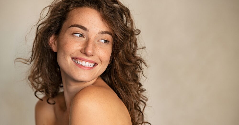 Woman with freckles and curly hair smiling against a neutral background
